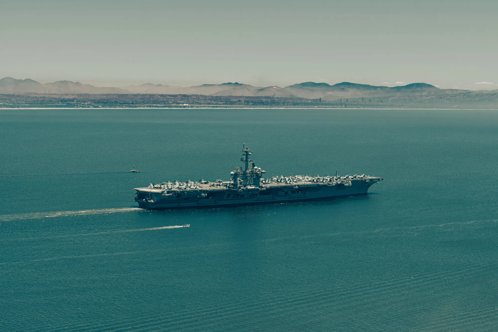 An aircraft carrier under way in coastal waters, its flight deck lined with aircraft, hills along the far shore